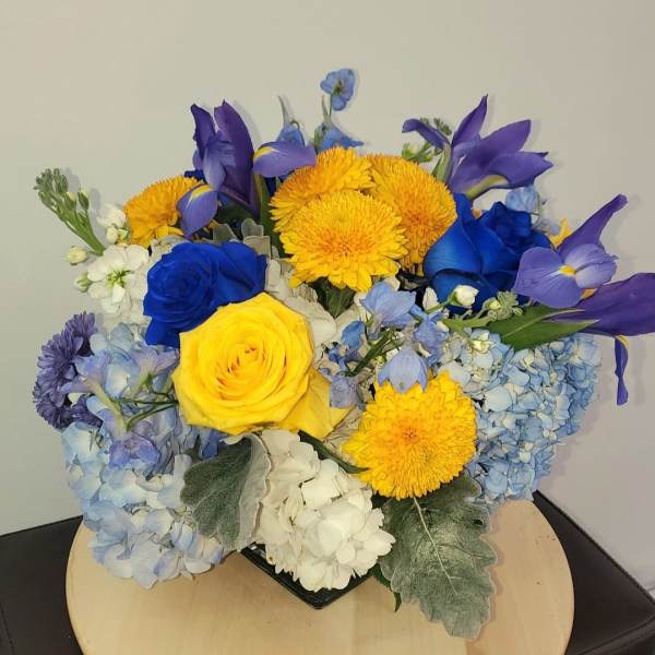 Blue and yellow floral arrangement with roses, hydrangeas, and daisies in a low container