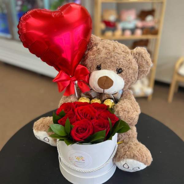 Teddy bear holding a red heart balloon beside red roses in a white box