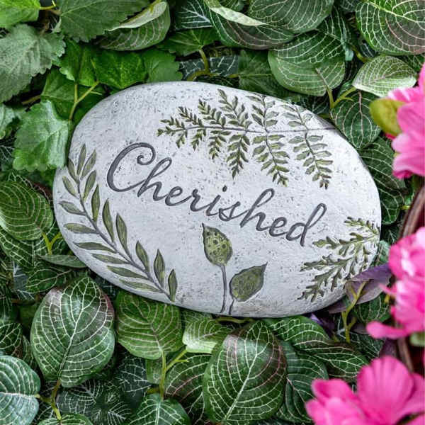 Decorative stone engraved with the word Cherished among green leaves and pink flowers