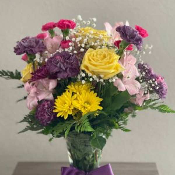 Mixed bouquet of yellow, pink, and purple flowers in a glass vase with a purple ribbon