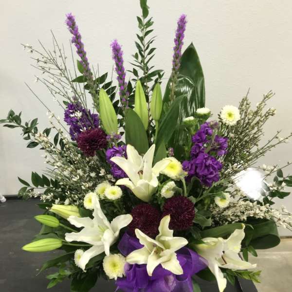 Tall floral arrangement with white lilies, purple blooms, and a purple ribbon in a vase