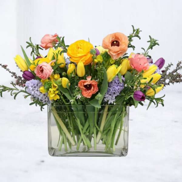 Low glass vase of yellow tulips, peach ranunculus, and purple hyacinths in a mixed spring arrangement