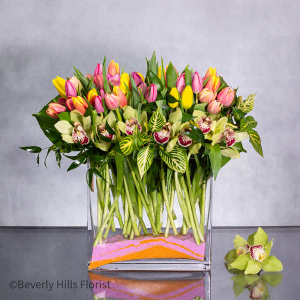 Rectangular vase of pink and yellow tulips with green orchids and variegated leaves over layered colored sand.
