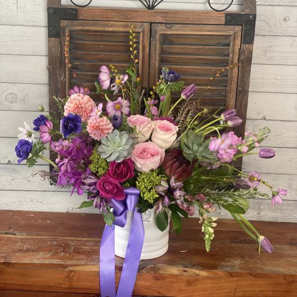 Mixed pink and purple flower arrangement in a white container with a ribbon