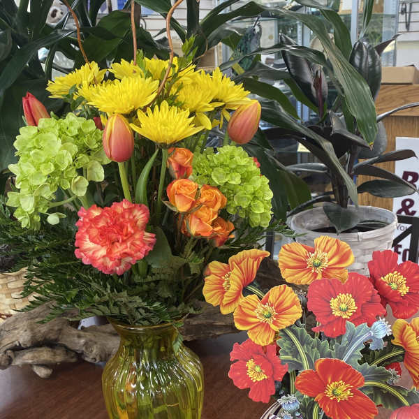 Bright mixed flower arrangement in a green glass vase beside a striped ceramic pot