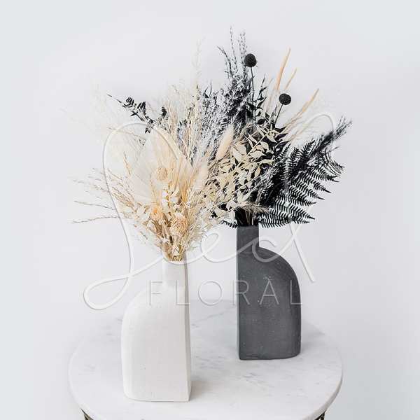 Two dried floral arrangements in white and black vases on a round table.