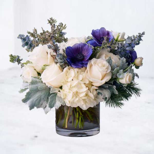 Low arrangement of white roses, hydrangeas, and purple anemones in a clear glass cylinder vase