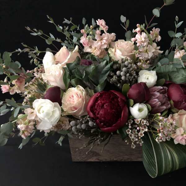 Low wooden box arrangement with blush roses, white ranunculus, and deep red blooms in a lush, elongated design