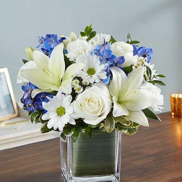 White lilies, roses, and blue flowers in a square glass vase
