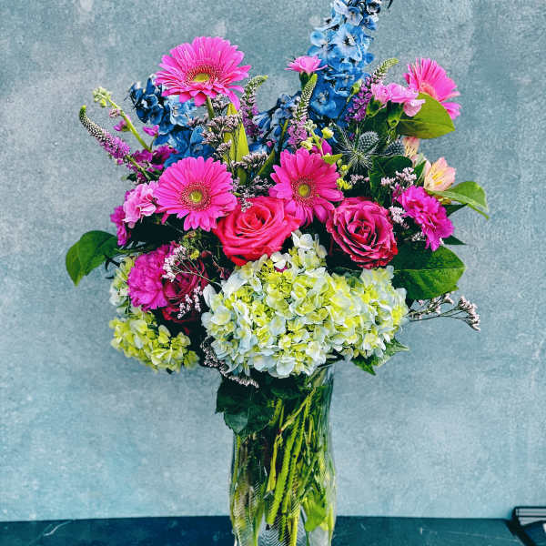 Bright mixed bouquet in a clear glass vase with pink gerberas, roses, hydrangeas, and blue delphinium.