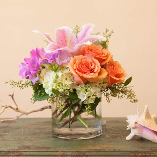 Pink lilies and orange roses in a clear glass vase with a seashell beside it.