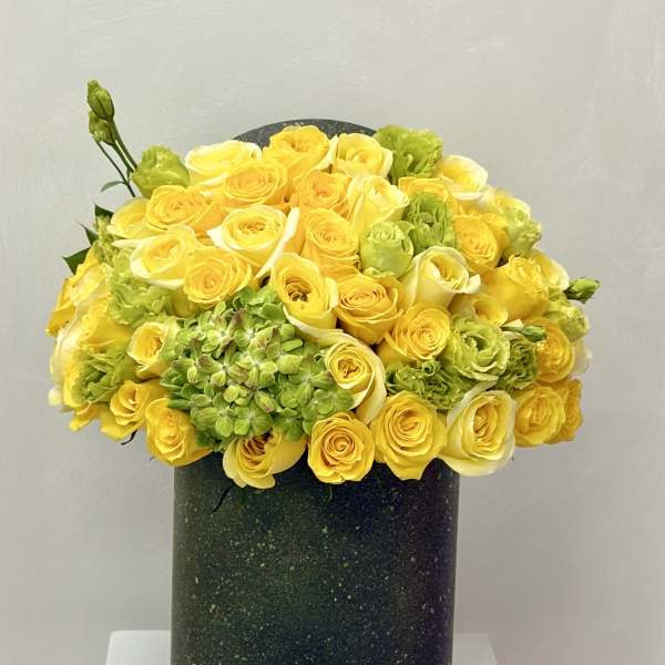 Dense arrangement of yellow and white roses with green blooms in a tall dark green cylinder vase