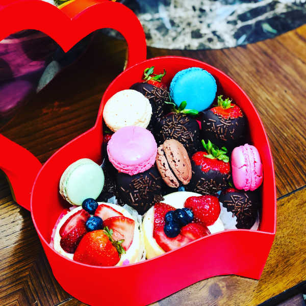 Heart-shaped red box filled with colorful macarons and chocolate-covered strawberries