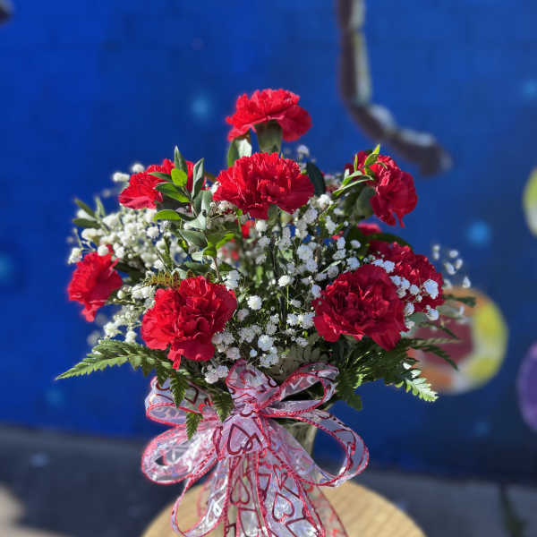 Red carnations in a glass vase with white filler flowers and a heart ribbon