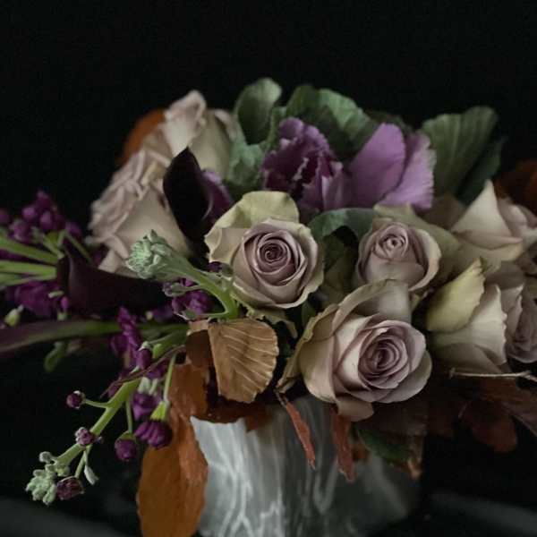 Moody arrangement of lavender roses, dark purple flowers, and kale in a white ceramic vase