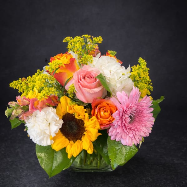 Mixed bouquet with roses, sunflowers, carnations, and pink daisies in a glass vase