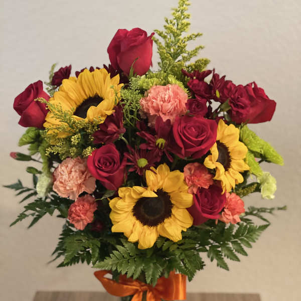 Bouquet of red roses, sunflowers, and carnations in a glass vase with orange ribbon