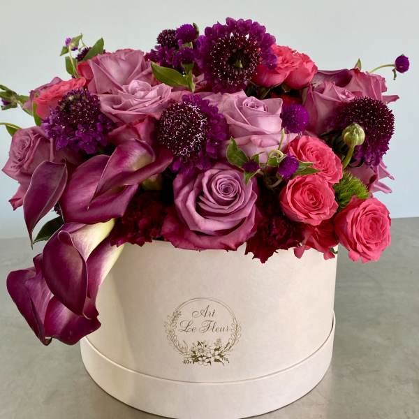 Pink and purple roses and calla lilies arranged densely in a round ivory hatbox container.