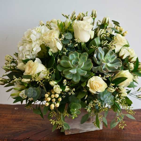 Compact arrangement of white roses, hydrangea, and succulents in a square vase.