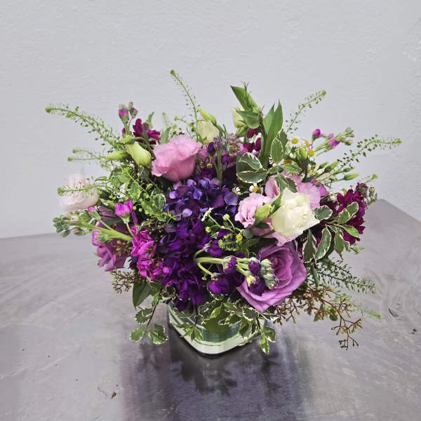 Low arrangement of purple and pink blooms with white accents in a clear glass cube vase