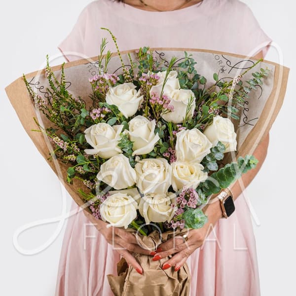 Hand-tied bouquet of white roses with small pink accent flowers wrapped in brown kraft paper.