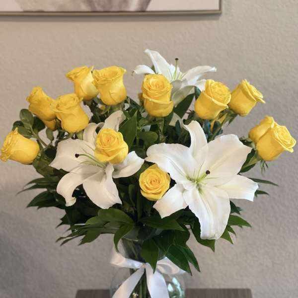 Yellow roses and white lilies arranged in a glass vase with a white ribbon.