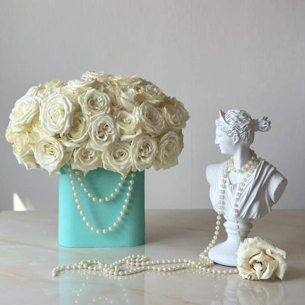 Cream rose arrangement in a turquoise box beside a white bust with pearl strands.