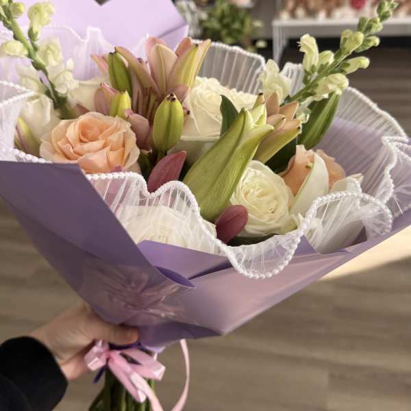 Hand-tied bouquet of white and peach roses with pink lilies, wrapped in lavender paper with ruffled white trim