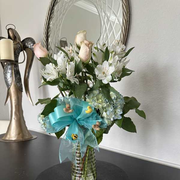 Vase arrangement of white alstroemeria, pale pink roses, and blue hydrangea with a teal bow and small baby-themed decorations.