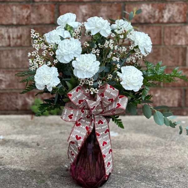 White carnations in a burgundy glass vase with a heart-patterned ribbon