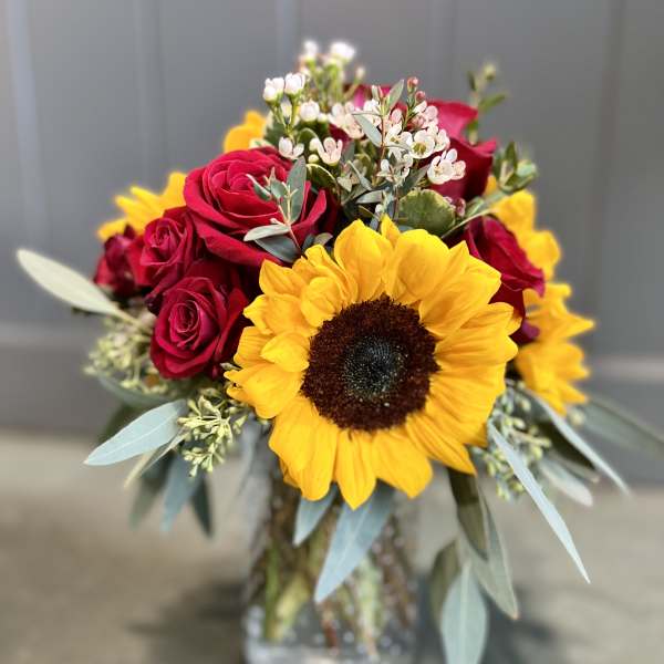 Arrangement of yellow sunflower, red roses, and small white blooms in a clear glass jar vase
