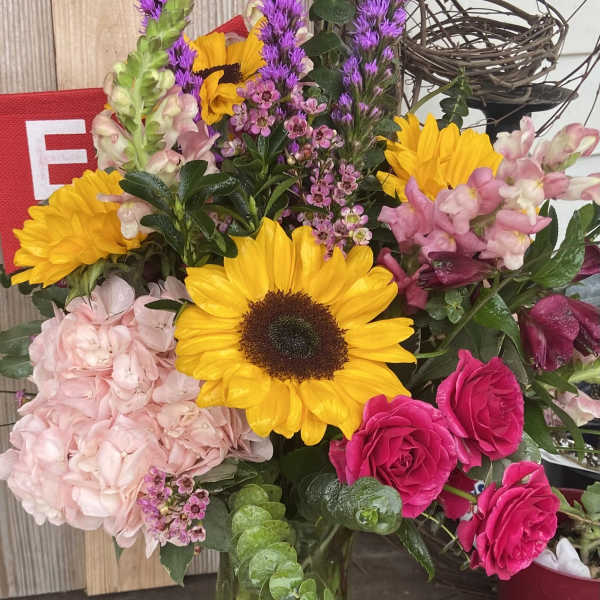 Mixed bouquet with sunflowers, pink roses, hydrangea, and purple spikes in a glass vase