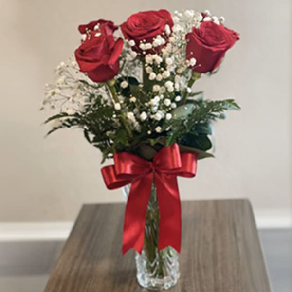 Three red roses in a clear vase with baby's breath and a red ribbon