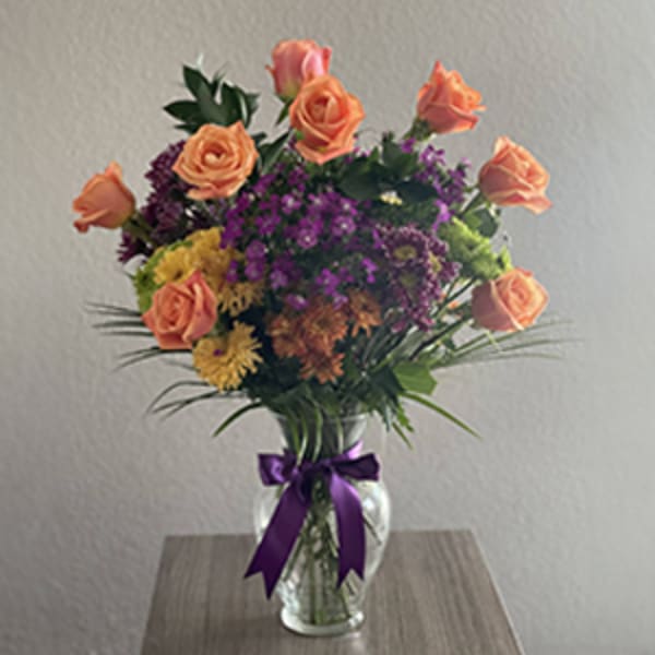 Bouquet of peach roses and purple filler flowers in a glass vase with a purple ribbon