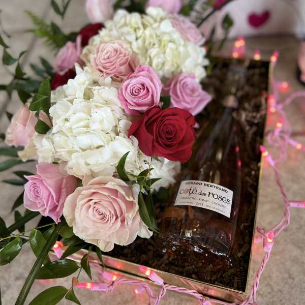 Pink and red roses with white hydrangeas in a gift box beside a wine bottle
