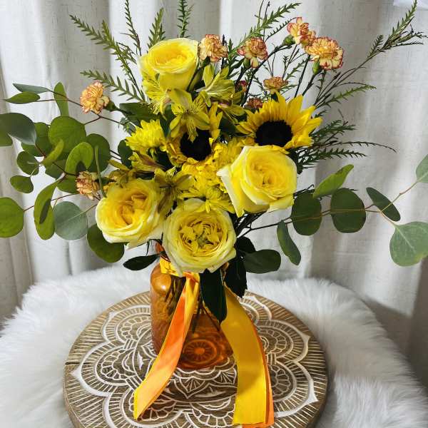 Yellow roses and sunflowers in an amber vase with orange ribbon