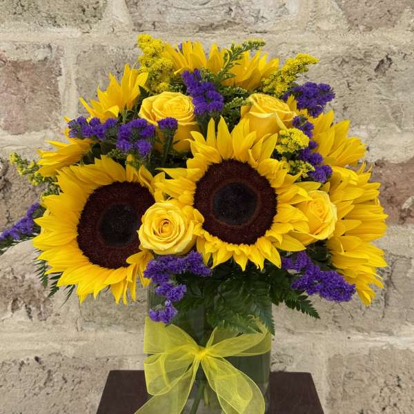 Sunflowers, yellow roses, and purple filler flowers in a glass vase with a yellow ribbon.