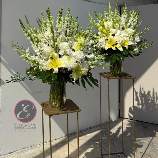 Two tall white and yellow floral arrangements in glass vases on stands