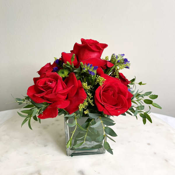 Red roses arranged in a square glass vase with greenery and small purple accents