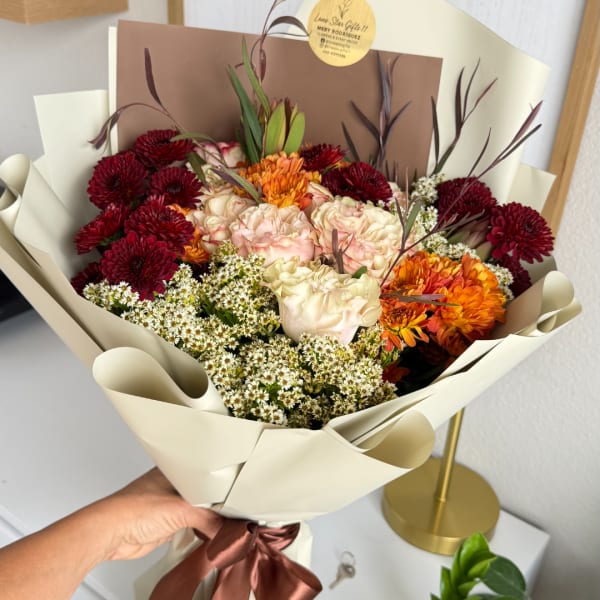 Handheld bouquet of burgundy, cream, and orange flowers wrapped in beige paper