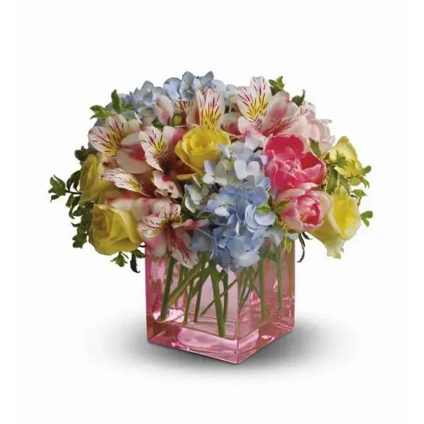 Low arrangement of yellow roses, blue hydrangeas and pink blooms in a pink glass cube vase