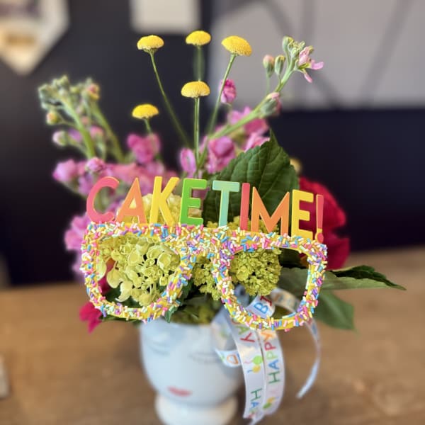 Flower arrangement in a white mug with a colorful "CAKE TIME!" topper