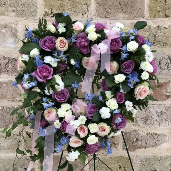 Heart-shaped floral wreath with purple, pink, and white roses on a stand