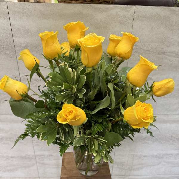 Tall arrangement of yellow roses in a clear glass vase on a wooden stand.