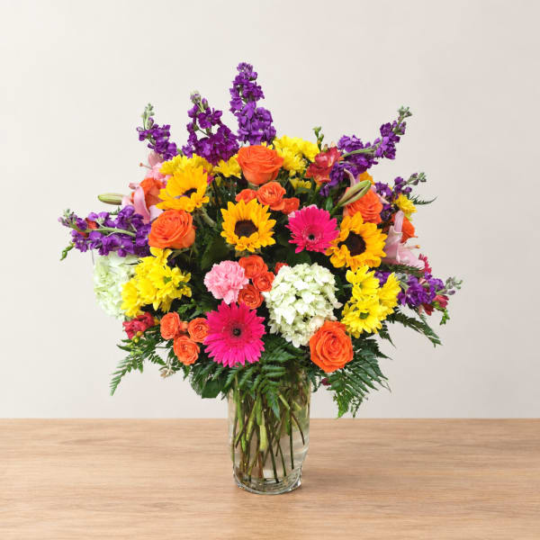 Colorful mixed bouquet in a clear glass vase with sunflowers, roses, and gerbera daisies