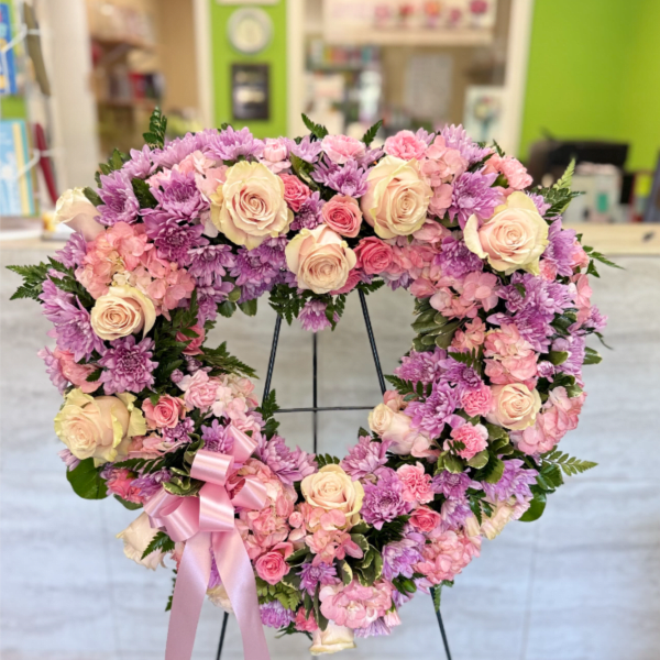 Heart-shaped standing wreath of pink and lavender flowers with a pink ribbon bow on a metal easel