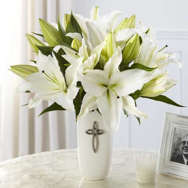 White lily arrangement in a tall white vase with a silver cross emblem on a light table.