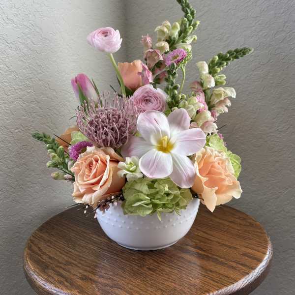 Pastel floral arrangement in a white bowl vase on a wooden table.