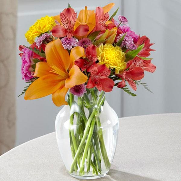 Mixed bouquet of orange, red, pink, and yellow flowers in a clear glass vase