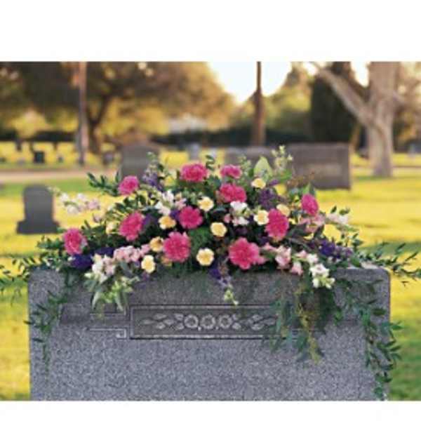 Graveside spray of pink, yellow, and white flowers draping over a gray headstone.